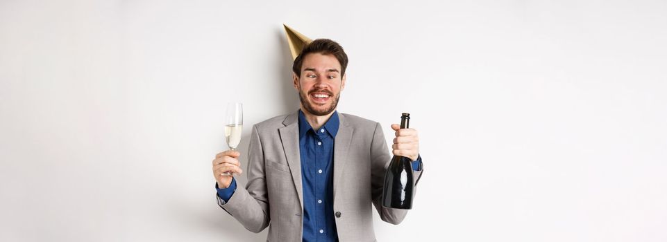 Celebration and holidays concept. Funny drunk guy in suit and birthday hat, squinting eyes and having fun at party, drinking champagne, white background.