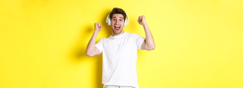 Excited handsome man dancing and singing along, listening music in headphones, standing over yellow background.