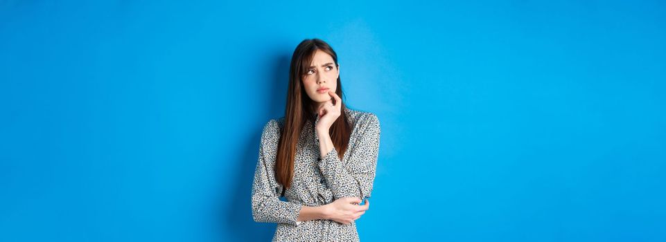 Thoughtful serious woman in dress looking away pensive, standing hesitant and unsure, thinking on blue background.