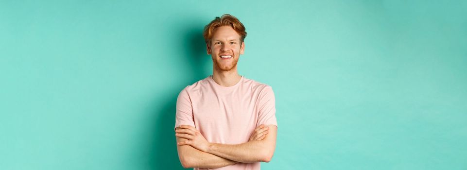 Portrait of friendly-looking young man with red hair and beard smiling and starng satisfied, holding hands crossed on chest, standing over turquoise background.