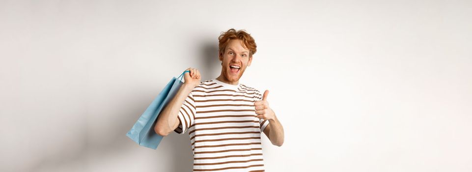 Happy young man with red hair, shopping in stores, showing thumbs-up and holding paper bag over shoulder, recommending shop, white background.
