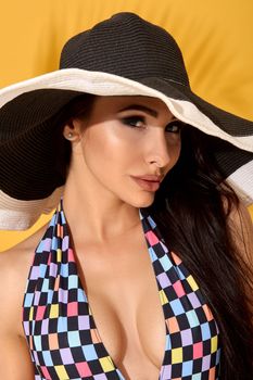 Photo of young beautiful brunette girl posing in the studio, standing in a black-and-white hat and checkered swimsuit. Yellow background