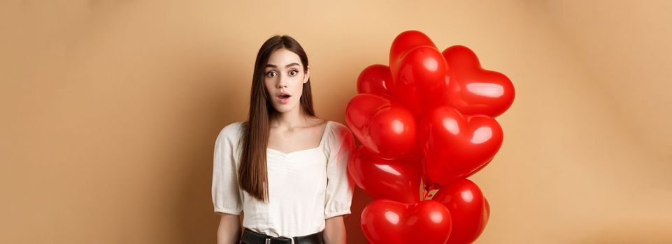 Surprised pretty girl standing near heart balloons and looking with awe at camera. Concept of Valentines day.