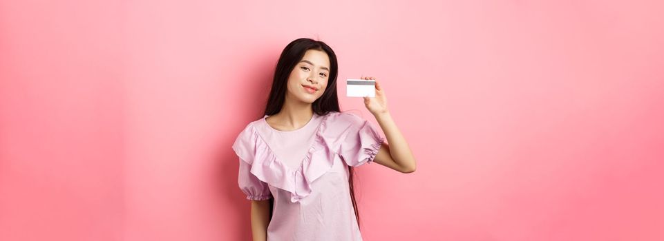 Shopping. Young korean girl in dress showing credit card, smiling satisfied at camera, standing on pink background.