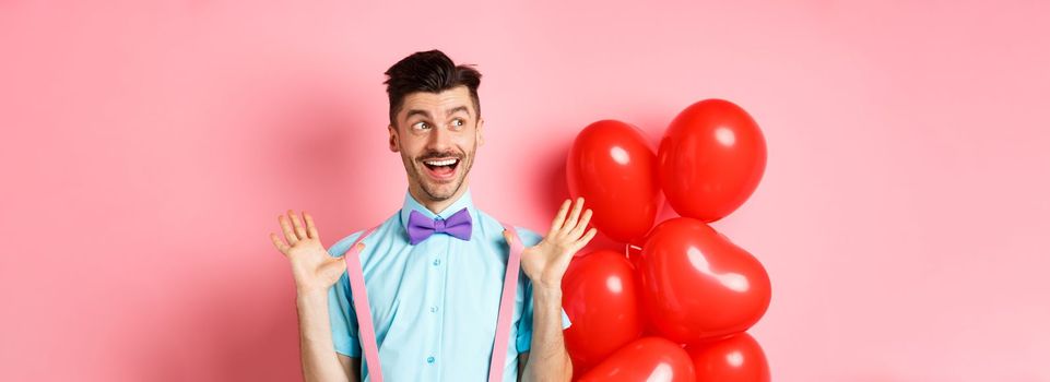Love and romance concept. Happy man screaming from fantastic news, shouting wow and smiling amused, checking out special offer on Valentines, standing near red hearts balloons.