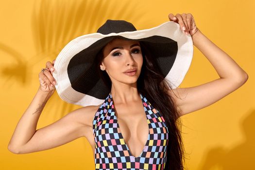 Photo of young beautiful brunette girl posing in the studio, standing in a black-and-white hat and checkered swimsuit. Yellow background