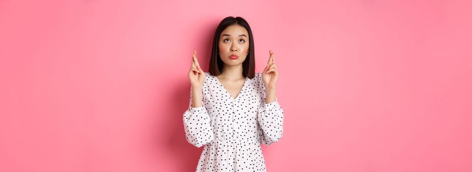 Cute asian woman making a wish, smiling happy and cross fingers for good luck, waiting over pink background.