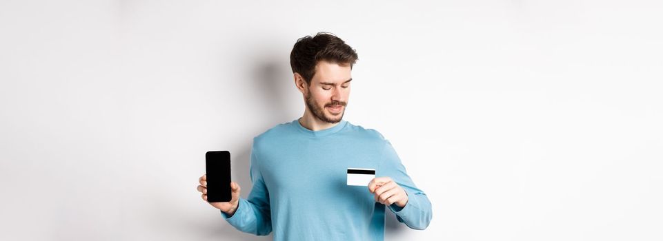 Handsome young man with beard, showing empty smartphone screen and looking at plastic credit card, standing over white background.