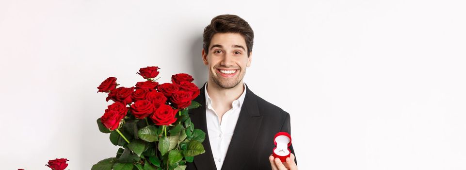 Close-up of attractive man in suit, holding bouquet of roses and engagement ring, making proposal, standing against white background.