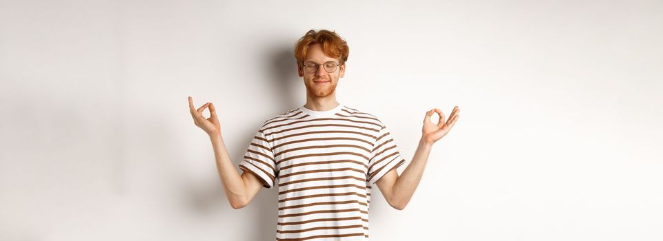 Calm and relaxed young man with red messy hair, spread hands sideways in mudra gesture and smiling, practice yoga or meditating, white background.