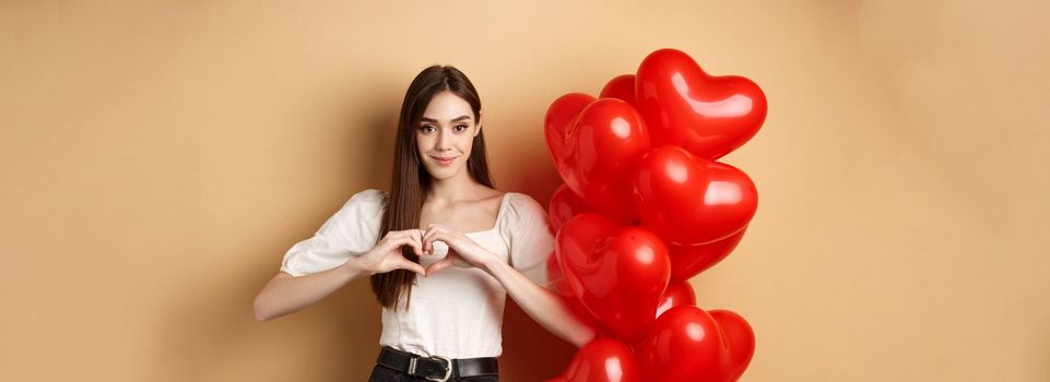 Image of beautiful young woman smiling and showing heart gesture, I love you sign, standing near romantic balloons on Valentines day, beige background.