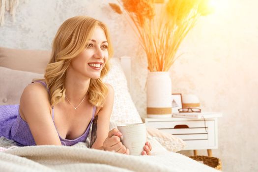 Young woman holding a mug with tea or coffee sitting on the bed in the bedroom. Near lie yarn and knitting needles. Horizontal photo. Freelance creative working and living concept.