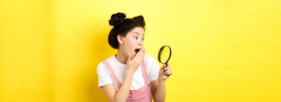 Excited silly asian girl gasping wondered, found something interesting, looking through magnifying glass, standing on yellow background.