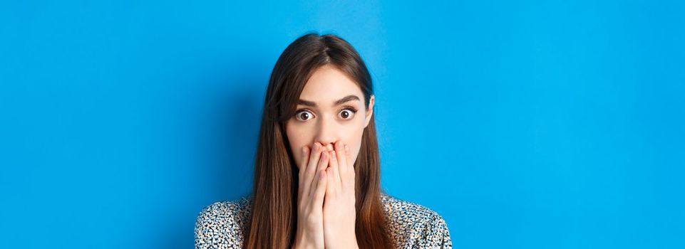 People emotions concept. Shocked young woman face, covering mouth with hands and gasping, look startled, standing on blue background.