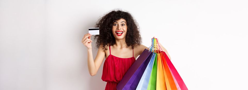 Stylish caucasian woman with curly hair and red dress, showing shopping bags and her plastic credit card, smiling amused, standing over white background.