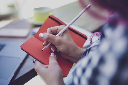 Positive female student with pink hair writing down funny story in notebook sitting in coffee shop with cup of cappuccino and laptop. Smiling female noting text in notepad