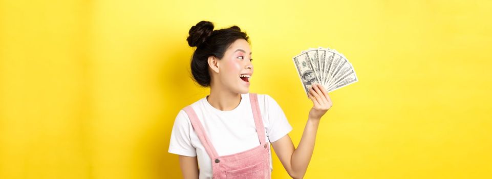 Shopping. Rich and stylish asian female model showing money, looking at dollar bills with happy face, standing on yellow background.