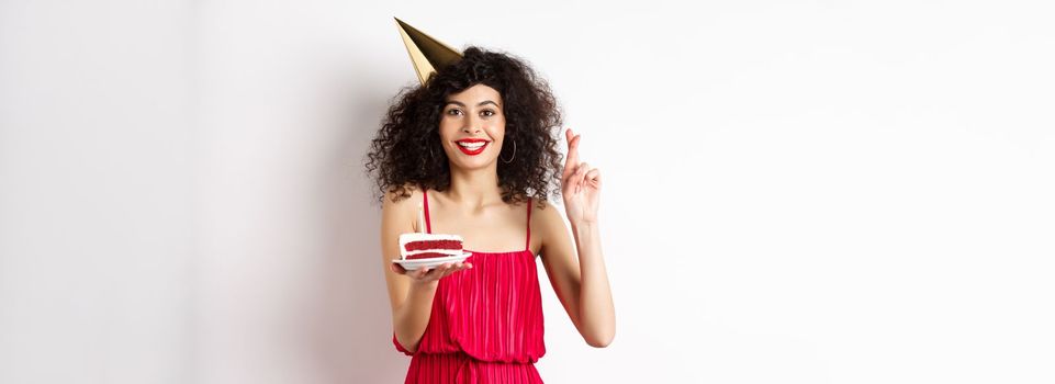 Excited birthday girl celebrating, making wish, holding cake and cross fingers good luck, standing happy on white background.