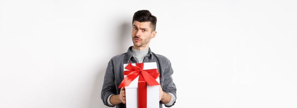 Surprised young man look with disbelief and hold surprise gift, raising eyebrow doubtful, being suspicious, standing on white background. Valentines day concept.