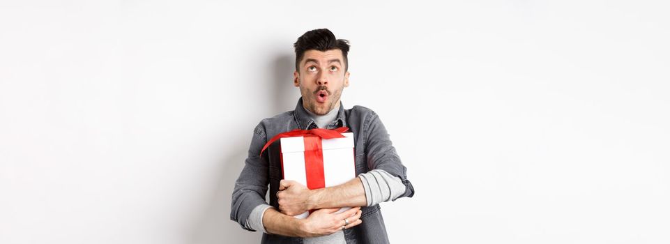 Surprised funny guy hugging big holiday gift and looking up, say wow amazed, checking out Valentines day special offer, standing on white background.