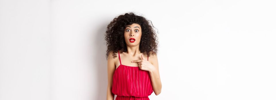 Surprised young woman in red dress pointing at herself, being confused and shocked, standing on white background. Copy space