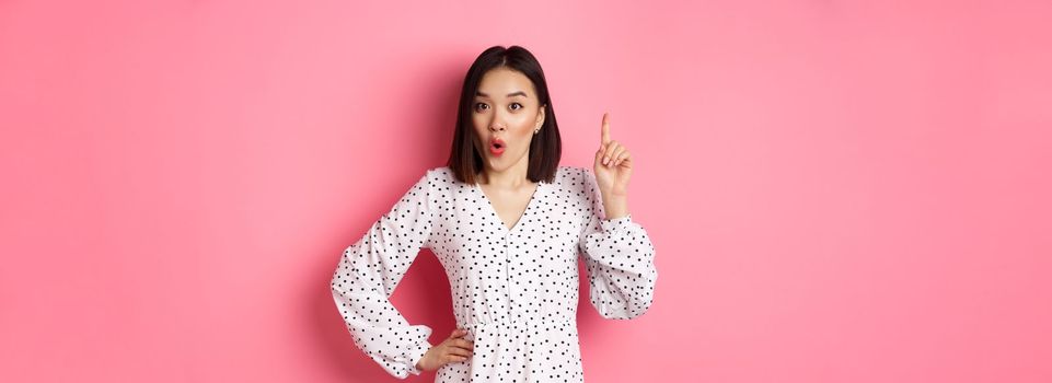 Excited asian girl having an idea, saying suggestion, raising finger and talking, standing over pink background.