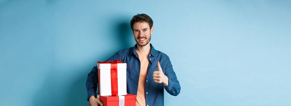 Smiling man holding romantic gifts and showing thumbs-up, celebrating Valentines day, buying presents for lover, standing over blue background.
