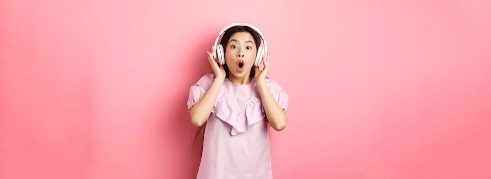 Excited asian girl hear awesome song, listen music in headphones and look amazed, standing on pink background.