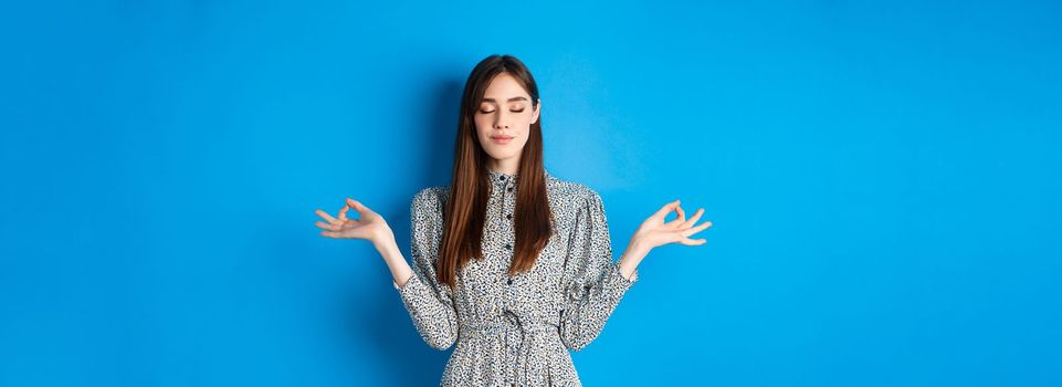 Mindful young woman in dress, close eyes and smile, meditating peaceful, standing with hands spread sideways with zen sign, blue background.