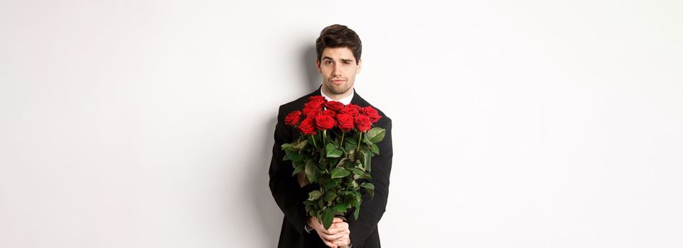 Image of handsome boyfriend in black suit, holding bouquet of red roses and smiling, being on a date, standing over white background.