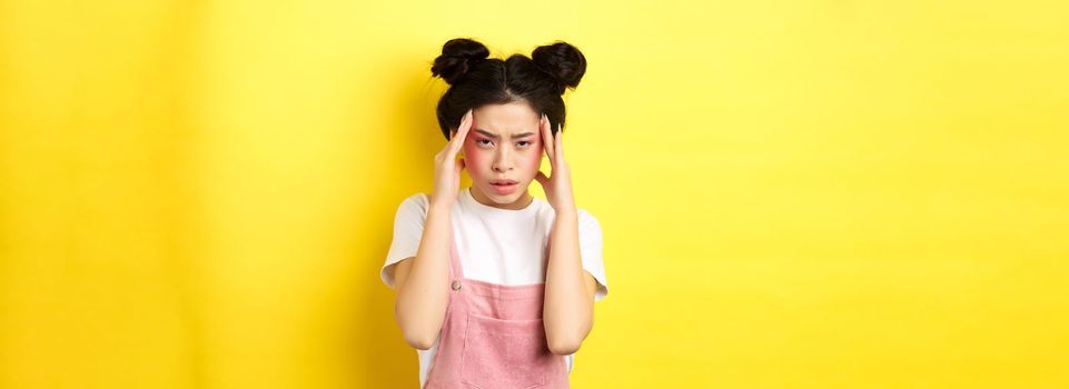 Image of stylish japanese woman with bright makeup, having headache, touching head and looking tired, have painful migraine, yellow background.