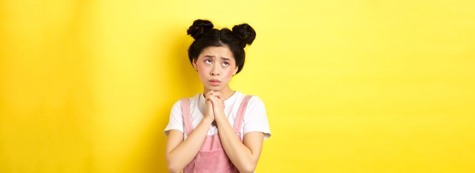 Sas asian girl holding hands in pray, pleading god, looking up and begging, standing on yellow background.