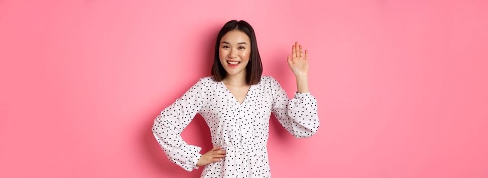 Beautiful asian woman in dress saying hello, waving hand to greet and say hi, smiling friendly at camera, standing over pink background.