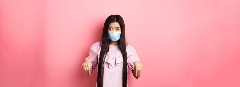 Healthy people and covid-19 pandemic concept. Sad asian woman pointing fingers down, frowning upset, standing against pink background.
