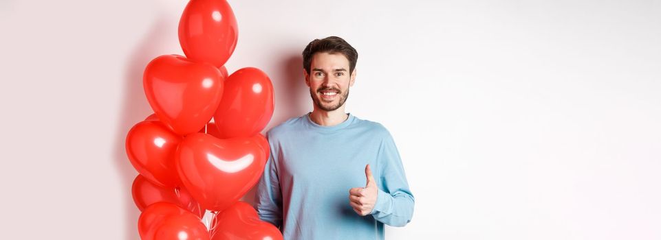 Valentines day and lovers concept. Happy young man showing thumbs up as standing with red hearts balloon, bring romantic gift on date, standing over white background.