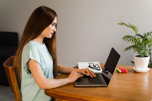 Candid girl with laptop is sitting on sofa smiling at successful work in IT company. Cheerful young woman programmer works remotely on laptop and try to meet deadline at home