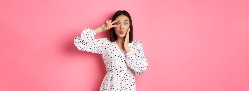 Coquettish asian woman in dress showing peace sign on eye, pucker lips for kiss, standing silly against pink background.