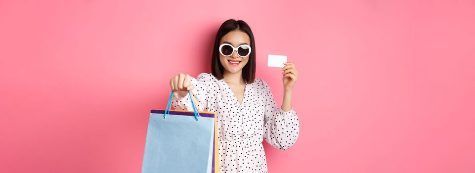 Beautiful asian woman in sunglasses going shopping, holding bags and showing credit card, standing over pink background.