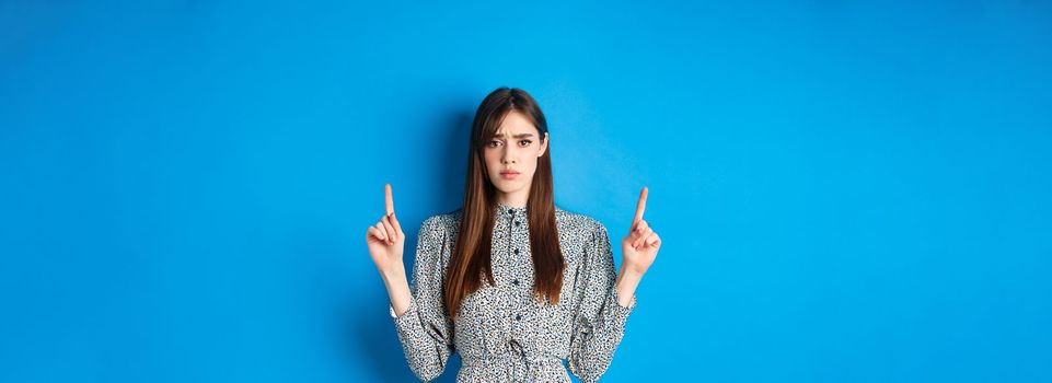 Hesitant and worried young woman frowning, pointing fingers up with sad face, standing on blue background.