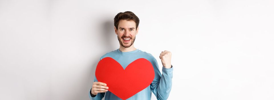 Cheerful man determined to find love on Valentines day, making fist pump success gesture and holding big red romantic heart, standing over white background.