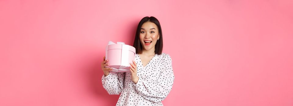 Valentines day. Happy asian woman receiving gift in cute box, smiling excited and thankful, standing in dress against pink background.