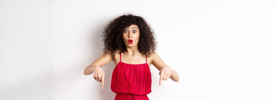 Impressed woman in red dress say wow, showing promo deal, pointing fingers down with amazed face, standing over white background.