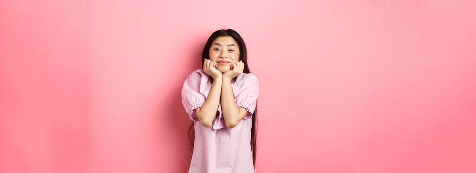 Cute and happy asian woman waiting for something with excitement, lean face on hands and watching lovely scene with admiration, standing against pink background.