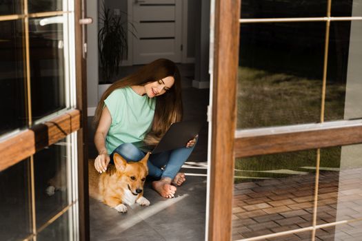 Attractive girl with laptop and Corgi dog sitting on the floor and chatting online with friends. Communication with family and having fun with dog together. Lifestyle with Welsh Corgi Pembroke