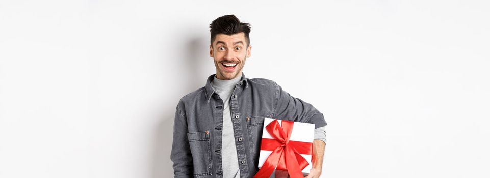 Happy Valentines day. Surprised and happy guy holding gift box and look at camera, smiling amazed, celebrating holiday, bring present on romantic date, white background.