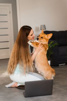Girl with laptop hug and kiss Welsh Corgi Pembroke dog at home. Young woman have a break and relax with her dog. Lifestyle with domestic pet