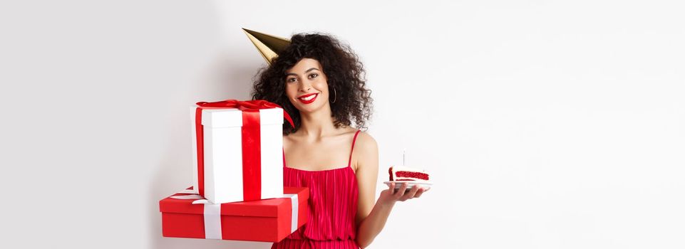 Holidays. Happy elegant woman in party hat and red dress, holding birthday cake and gifts, celebrating b-day, standing over white background.