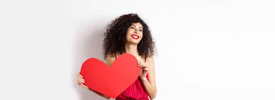 Romantic woman in dress and makeup, looking aside with dreamy face and love, showing big red valentines heart card, standing on white background.