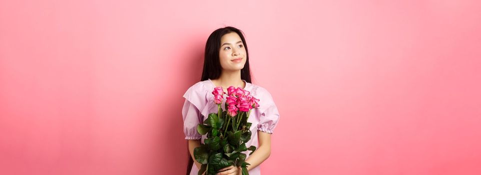 Asian teenage girl in cute dress looking romantic at empty space logo, holding valentines day flowers gift, receive boquet of roses from lover, standing on pink background.