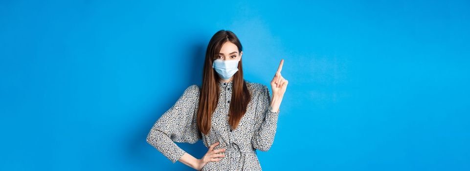 Healthy people and covid-19 pandemic concept. Serious woman in medical mask pointing at upper left corner, standing on blue background.